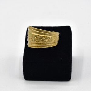 May include: A gold-colored ring with a floral design. The ring is displayed on a black velvet surface against a white background. The ring's band features embossed flowers and leaves.