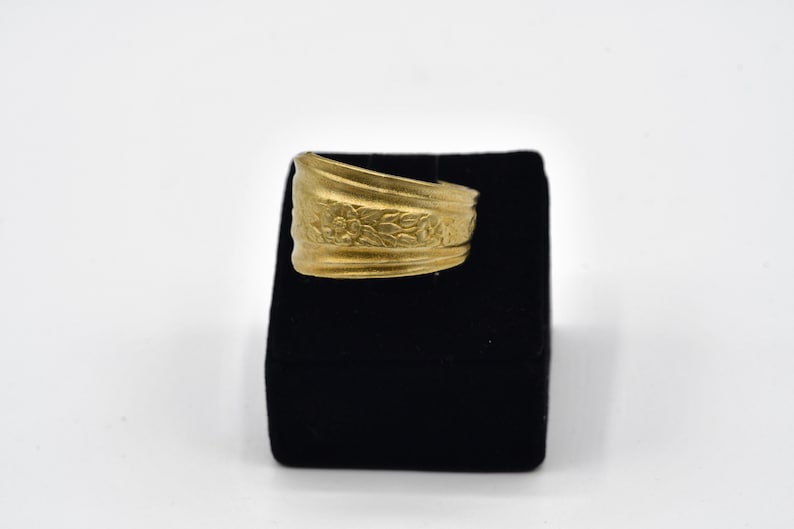 May include: A gold-colored ring with a floral design. The ring is displayed on a black velvet surface against a white background. The ring's band features embossed flowers and leaves.