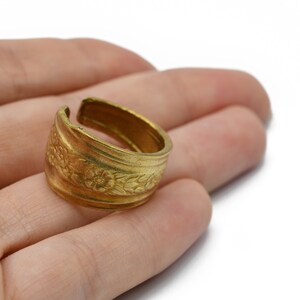 May include: A gold-colored adjustable ring with a floral pattern. The ring is open at the top and features detailed embossed flowers and leaves around the band. The ring is likely made of brass or a similar metal.