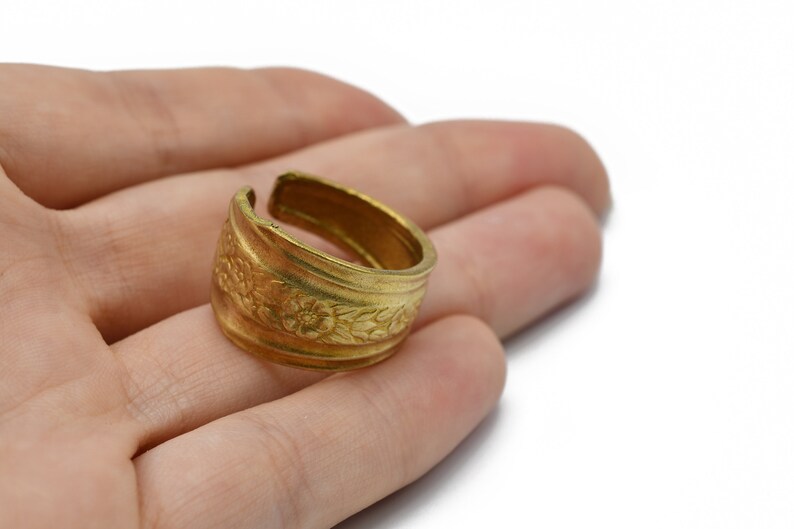 May include: A gold-colored adjustable ring with a floral pattern. The ring is open at the top and features detailed embossed flowers and leaves around the band. The ring is likely made of brass or a similar metal.