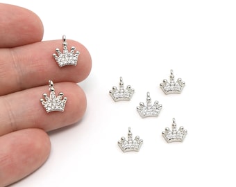 May include: Small silver-colored crown charms with clear, sparkling stones. The charms have a loop at the top for attaching to jewelry. Several charms are scattered on a white surface, with some held by a hand.