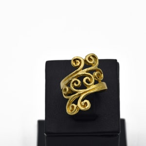 May include: A gold-colored ring with an open design featuring swirling, scroll-like patterns. The ring is displayed on a black stand against a white background. The jewelry piece has a bright, polished finish.