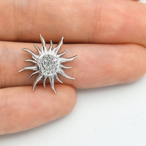 May include: A silver-colored sun-shaped pendant with a central cluster of small, sparkling stones. The pendant has pointed rays extending outwards, creating a sunburst design. The jewelry is held in a hand against a white background.