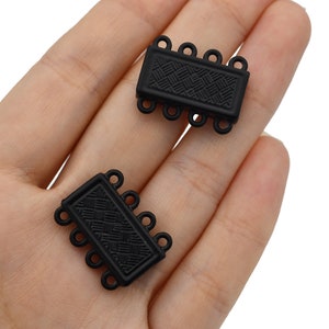 May include: Two black rectangular jewelry clasps with a woven pattern are displayed. Each clasp has four small loops on each end for attaching to jewelry. The clasps are made of a matte black material.