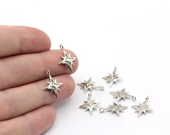 Puede incluir: Una colección de dijes de estrella plateados con un diseño en capas. Cada dije tiene un pequeño lazo en la parte superior para sujetarlo a la joyería. Los dijes están dispersos sobre una superficie blanca.