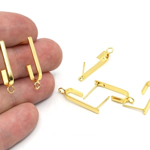 May include: Gold-colored earring components with a post and a curved, rectangular design. The earrings have a small loop at the bottom for attaching charms or other decorative elements. Several pieces are scattered on a white surface.