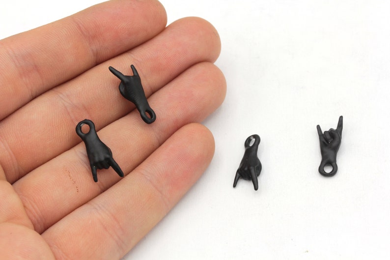 May include: Four small, black charms shaped like hands making the 'rock on' hand gesture. Each charm has a small loop at the top for attaching to jewelry or other crafts. The charms are displayed on a light background.