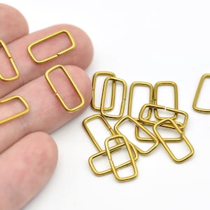 Könnte beinhalten: Eine Sammlung kleiner, rechteckiger, goldfarbener Metallringe. Die Ringe sind auf einer Seite offen und scheinen neu zu sein. Sie sind auf einer weißen Oberfläche verstreut, einige werden in einer Hand gehalten. Dies sind wahrscheinlich Schmuckzubehör.