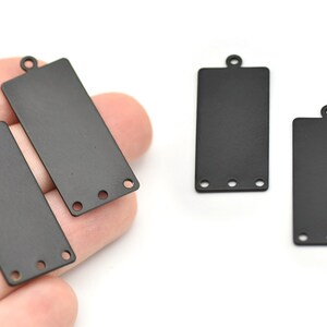 Puede incluir: Componentes de joyería rectangulares de metal negro con un lazo en la parte superior y tres agujeros en la parte inferior. Estos componentes son ideales para crear pendientes, colgantes u otros diseños de joyería. Fondo blanco.