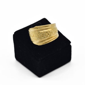 May include: A gold-colored ring with a floral pattern. The ring is displayed on a black velvet box against a white background. The ring's design features embossed flowers and leaves, creating a textured surface.