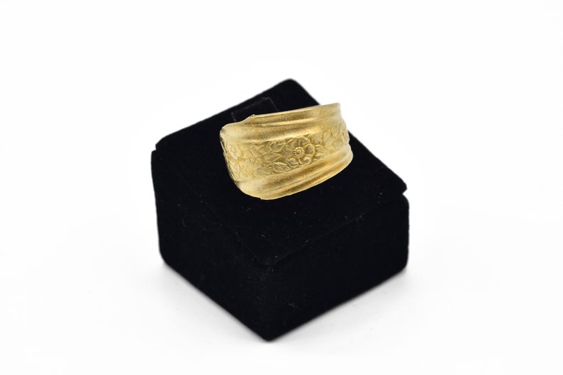 May include: A gold-colored ring with a floral pattern. The ring is displayed on a black velvet box against a white background. The ring's design features embossed flowers and leaves, creating a textured surface.