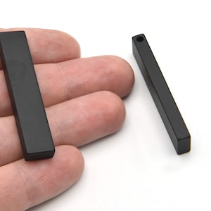 Könnte beinhalten: Zwei schwarze, rechteckige Anhänger mit matter Oberfläche. Einer wird in der Hand gehalten, der andere liegt separat. Jeder hat ein kleines Loch oben, was darauf hindeutet, dass sie für die Schmuckherstellung bestimmt sind. Sie sind etwa 2,5 cm lang.