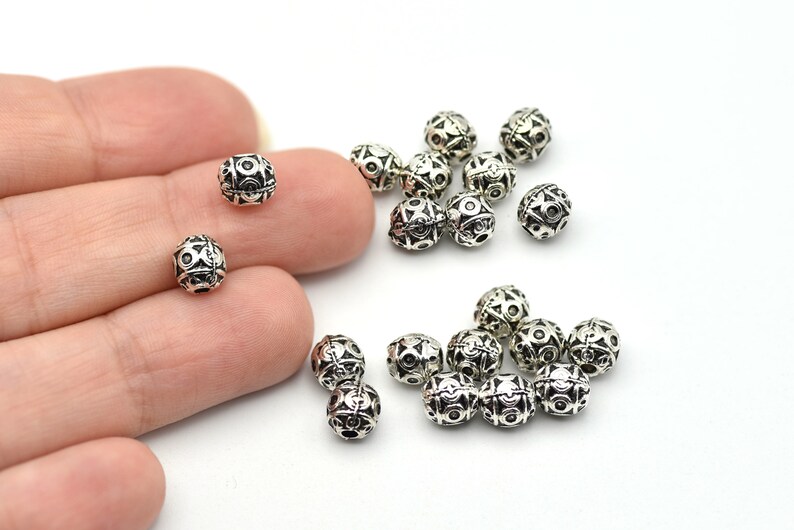 May include: Silver-colored metal beads with intricate geometric patterns. The beads are small, round, and have a detailed design of circles and lines. Some beads are held in a hand, while others are scattered on a white surface. These beads are suitable for jewelry making.