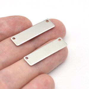 May include: Two rectangular, silver-coloured metal jewellery tags with rounded corners and two small holes at each end. The tags are blank and smooth, suitable for engraving or stamping. They are held in a person's hand against a white background.