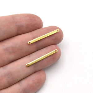 May include: Two small, rectangular gold-colored metal bars with a hole at each end. The bars are held in a person's hand against a white background. These components are likely for jewelry making.