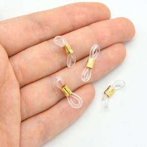 K&ouml;nnte beinhalten: Vier transparente Brillenhalterungen mit goldfarbenen Metallakzenten. Die Halterungen bestehen aus einem flexiblen, transparenten Material und sind zur Befestigung an den B&uuml;geln von Brillen vorgesehen. Die goldenen Akzente verleihen dem funktionellen Accessoire einen Hauch von Eleganz.