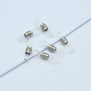 May include: A collection of six clear eyeglass retainers with silver-colored metal accents. The retainers are designed to loop around the earpieces of glasses, providing a secure fit. The background is a light blue surface.