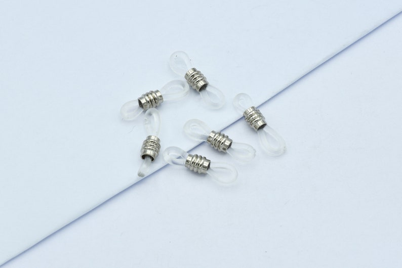 May include: A collection of six clear eyeglass retainers with silver-colored metal accents. The retainers are designed to loop around the earpieces of glasses, providing a secure fit. The background is a light blue surface.
