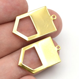 Könnte beinhalten: Zwei goldfarbene Metallanhänger. Jeder Anhänger hat eine rechteckige Form mit einem geometrischen Ausschnitt und einer kleinen Schlaufe oben zum Befestigen an einer Kette oder einer anderen Schmuckkomponente. Die Anhänger sind glänzend und reflektierend.