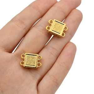 May include: Two gold-colored square jewelry clasps with a woven pattern on the front. Each clasp has two loops on each side. The clasps are held in a person's hand against a white background.
