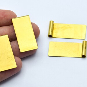 May include: Several rectangular, gold-colored metal jewelry findings. Two have a rolled edge, and the others are flat. The findings are displayed on a white background.