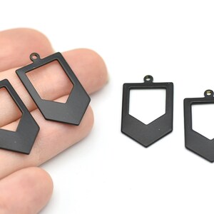 Może przedstawiać: Czarne metalowe zawieszki z geometrycznym wzorem. Zawieszki mają prostokątny kształt z wyciętym środkiem i strzałką skierowaną w dół. Każda zawieszka ma małą pętlę na górze do mocowania biżuterii.