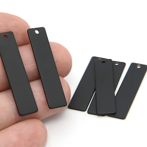 May include: Several matte black rectangular metal tags with a hole at the top for jewelry making or crafting. Some tags are held in a hand, while others are scattered on a white surface.