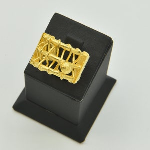 May include: A gold-colored ring with a rectangular cage-like design. The ring features a central sphere and intricate bar detailing. It is displayed on a black stand against a white background. The ring is a piece of jewelry.