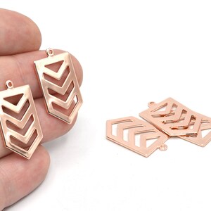 May include: Rose gold-colored metal jewelry findings. The rectangular charms feature a cut-out chevron design and a small loop at the top for attaching to other jewelry components. Several pieces are visible.