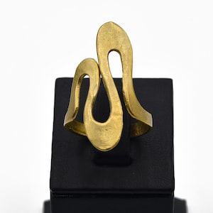 May include: A gold-colored ring with an abstract design. The ring features a flowing, openwork pattern with curved lines and a central teardrop shape. The ring is displayed on a black stand against a white background.