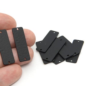 May include: Several matte black rectangular metal tags with textured surfaces. Each tag has a hole at the top and bottom. The tags are likely used for jewelry making or crafting projects.
