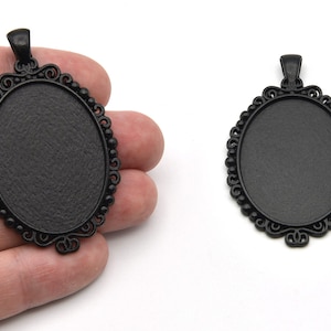 May include: Two black oval pendant trays with ornate filigree edges. The trays have a textured surface and a loop at the top for a chain. The pendants are displayed against a white background.