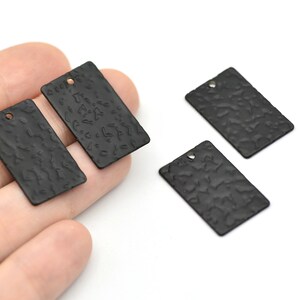 May include: Black rectangular metal tags with a textured surface and a small hole at the top. The tags are shown against a white background. These tags are suitable for jewelry making and crafting.