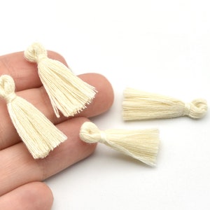 May include: Close-up of several small, cream-colored tassels. Each tassel is made of tightly bundled threads, with a looped top and a fringed bottom. The tassels are likely for crafting or embellishment.