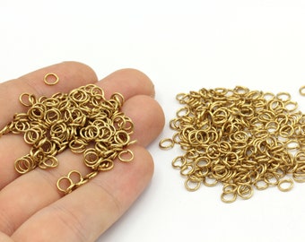 Puede incluir: Una colección de pequeños anillos de salto dorados, perfectos para la fabricación de joyas o manualidades. Los anillos son redondos y parecen ser de metal. Algunos anillos se sostienen en una mano, otros están dispersos.