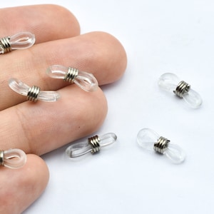 May include: Clear, oval-shaped eyeglass retainers with silver metal coils. These small, transparent accessories are designed to secure eyeglasses. Several retainers are scattered on a white surface.