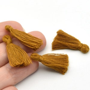 May include: Four small, mustard-colored tassels. Each tassel is made of many strands of thread, gathered at the top and fanning out at the bottom. The tassels are on a white background.