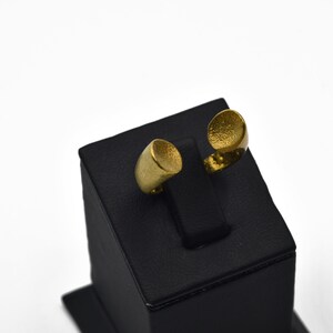 Op de afbeelding: Twee goudkleurige ringen tentoongesteld op een zwarte standaard. Eén ring heeft een cilindrische vorm, de andere heeft een getextureerd, open ontwerp. De ringen worden gepresenteerd tegen een witte achtergrond, wat hun metallic afwerking benadrukt.
