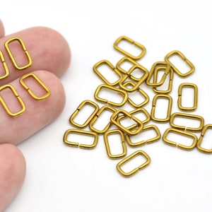 May include: A collection of small, rectangular, gold-colored metal rings. Some rings are grouped together, while others are scattered on a white background. These hardware findings are likely used for jewelry making or crafting.