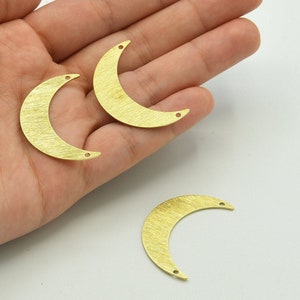 Puede incluir: Tres colgantes dorados en forma de media luna con superficie texturizada. Cada colgante tiene dos pequeños agujeros para ensartar. Los colgantes están dispuestos sobre un fondo claro.