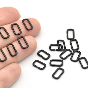May include: A collection of small, matte black rectangular metal rings, likely for crafting or jewelry making. Some rings are held in a hand, while others are scattered on a white surface. The rings have a simple, open design.