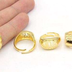 May include: Gold-colored adjustable ring with the word "AMOUR" cut out in the center. The ring has an oval face and a smooth, polished finish. The ring is shown in multiple views.