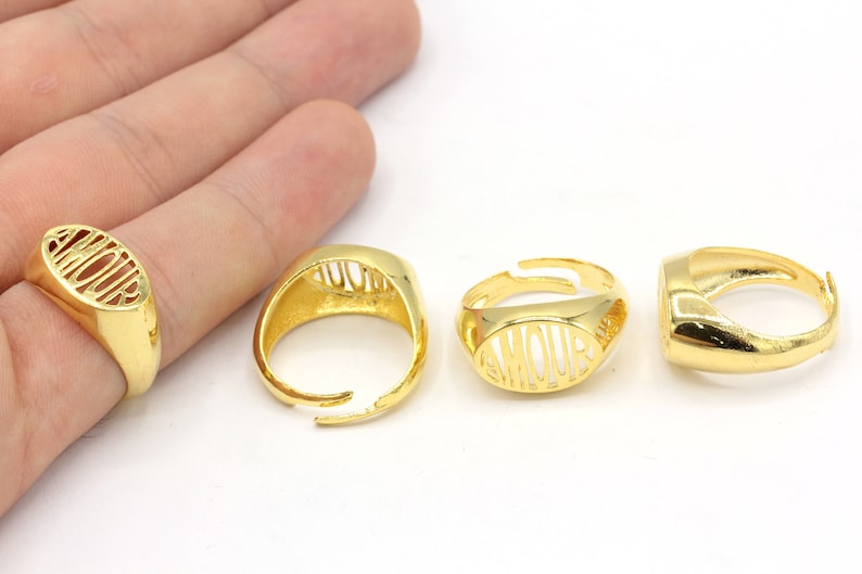 May include: Gold-colored adjustable ring with the word "AMOUR" cut out in the center. The ring has an oval face and a smooth, polished finish. The ring is shown in multiple views.