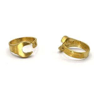 May include: Two gold-colored rings shaped like miniature wrenches. The rings are made of a shiny, metallic material and are set against a white background. The wrench design is clearly visible, with the open-ended jaws and the handle.