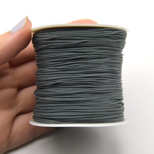 May include: A spool of dark gray elastic cord. The cord is tightly wound around a white plastic spool. The image is a close-up shot, highlighting the texture and color of the elastic material. Ideal for jewelry making and crafts.