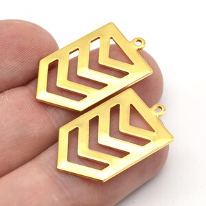 Könnte beinhalten: Zwei goldfarbene geometrische Anhänger mit rechteckiger Form und Zickzack-Design. Jeder Anhänger hat oben eine kleine Schlaufe zum Befestigen an Schmuck. Die Anhänger werden vor weißem Hintergrund gezeigt.