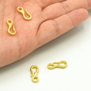May include: Four small, gold-colored jewelry clasps. Each clasp has a hook shape with a looped end and a wrapped design on the other end. The clasps are shown on a light background.