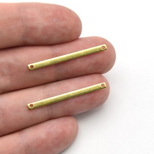 May include: Two small, rectangular, brass-colored metal bars with a hole at each end. The bars are held between fingers against a white background. These are likely jewelry-making components.