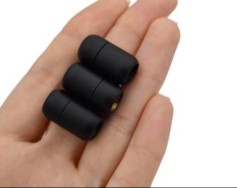 May include: Three black cylindrical cord end caps with a matte finish are shown. The caps are held in a hand, and the ends have a small gold-coloured interior. These caps are likely used for jewellery making or crafting.