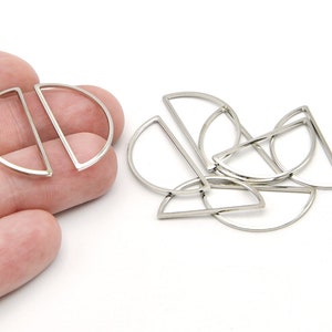 Pode incluir: Berloques geométricos de metal prateado. Os berloques têm a forma de um semicírculo com uma linha reta que corta o lado curvo. Vários berloques estão espalhados sobre um fundo branco.
